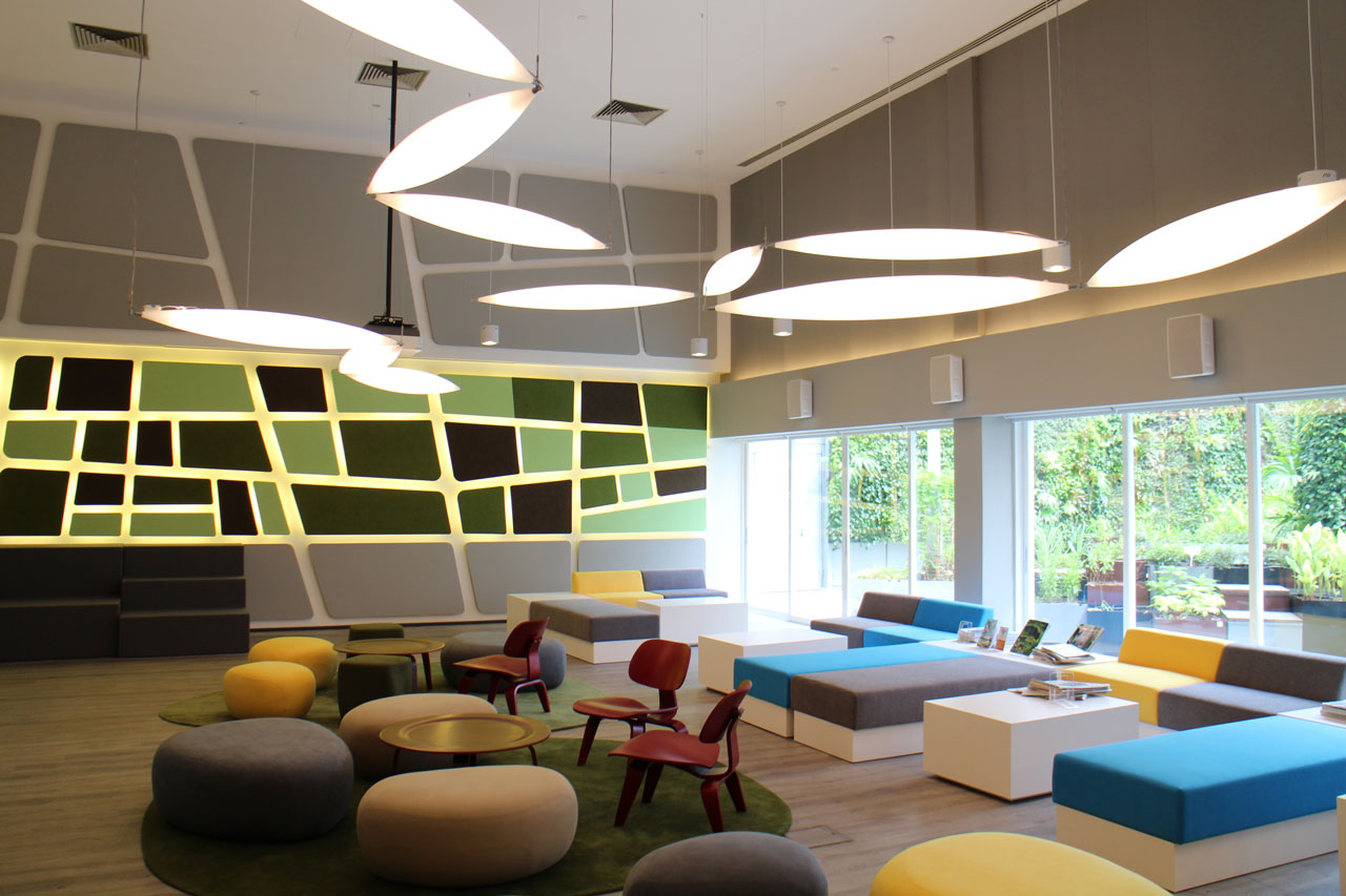 A modern lounge with geometric patterned walls, large glowing pendant lights, colorful round ottomans, red chairs, and long white benches. Floor-to-ceiling windows reveal lush greenery outside.