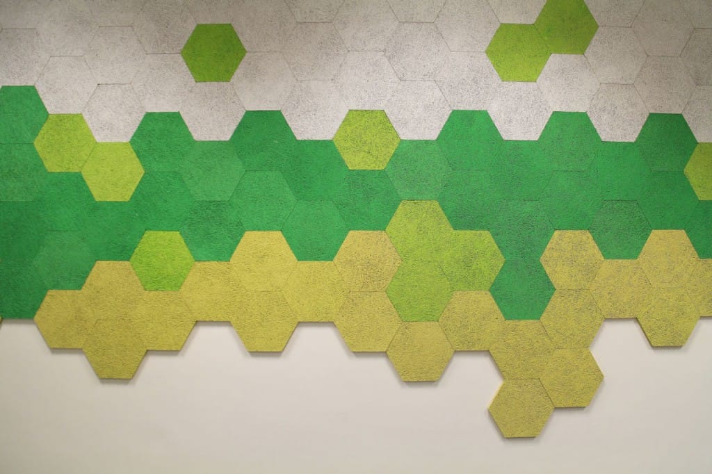 A wall covered with hexagonal tiles arranged in a honeycomb pattern, featuring a gradient of colors from white at the top, transitioning through various shades of green, to yellow at the bottom.