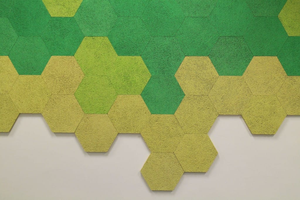 Hexagonal panels in varying shades of green and yellow arranged in a honeycomb pattern on a white wall, creating a gradient effect from darker green at the top to yellow at the bottom.
