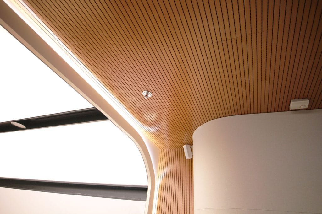 Modern interior ceiling with light wood slats, integrated lighting, and sleek white curved walls. Large windows with black frames allow natural light to enter, creating a bright and minimalistic atmosphere.
