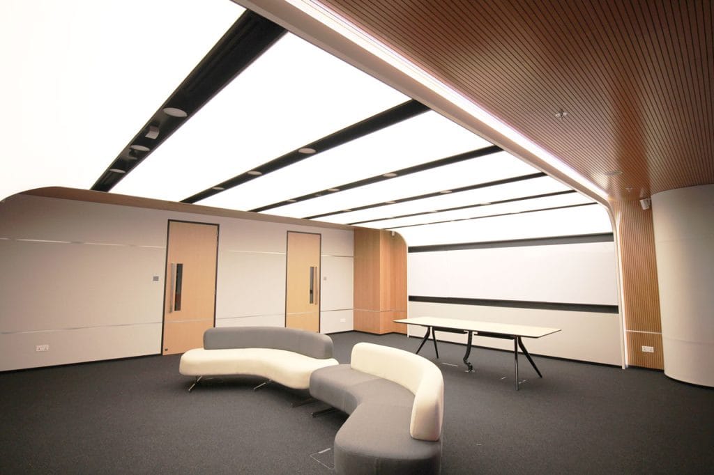 A modern, minimalistic room with curved light panels on the ceiling, light wood doors and walls, a curvy gray and white sectional sofa, and a long table on a black carpet floor.