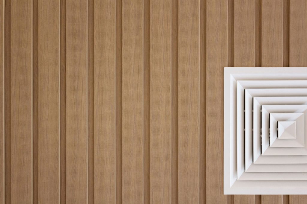 A white air vent with a square, layered design is mounted on a light brown, vertically grooved wall or ceiling. The vent is positioned in the upper left corner of the image.
