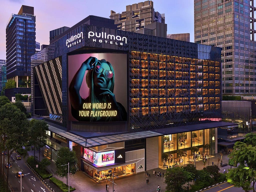 A modern Pullman Hotel building at dusk features a large billboard of a woman in sunglasses with the text OUR WORLD IS YOUR PLAYGROUND. The street below is busy, with shops including Adidas at ground level.