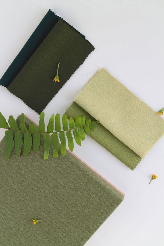 Three green fabric swatches are arranged on a white surface with a fern leaf and small yellow flowers scattered around, creating a natural, earthy aesthetic.