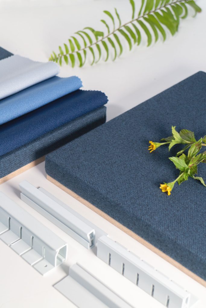 Fabric swatches in shades of blue, a textured blue panel, pieces of white hardware, and green foliage are arranged on a white surface.
