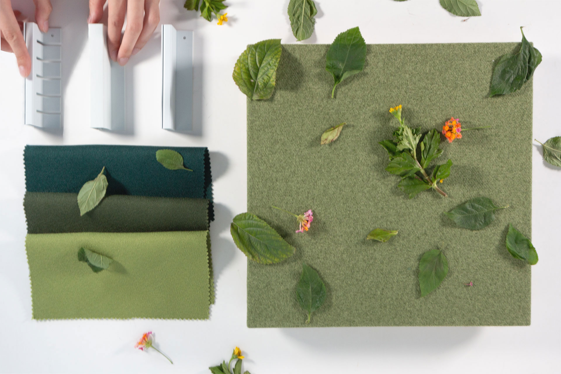 A hand arranges objects beside three green fabric swatches and a green textured board, both decorated with scattered leaves and small colorful flowers on a white surface.