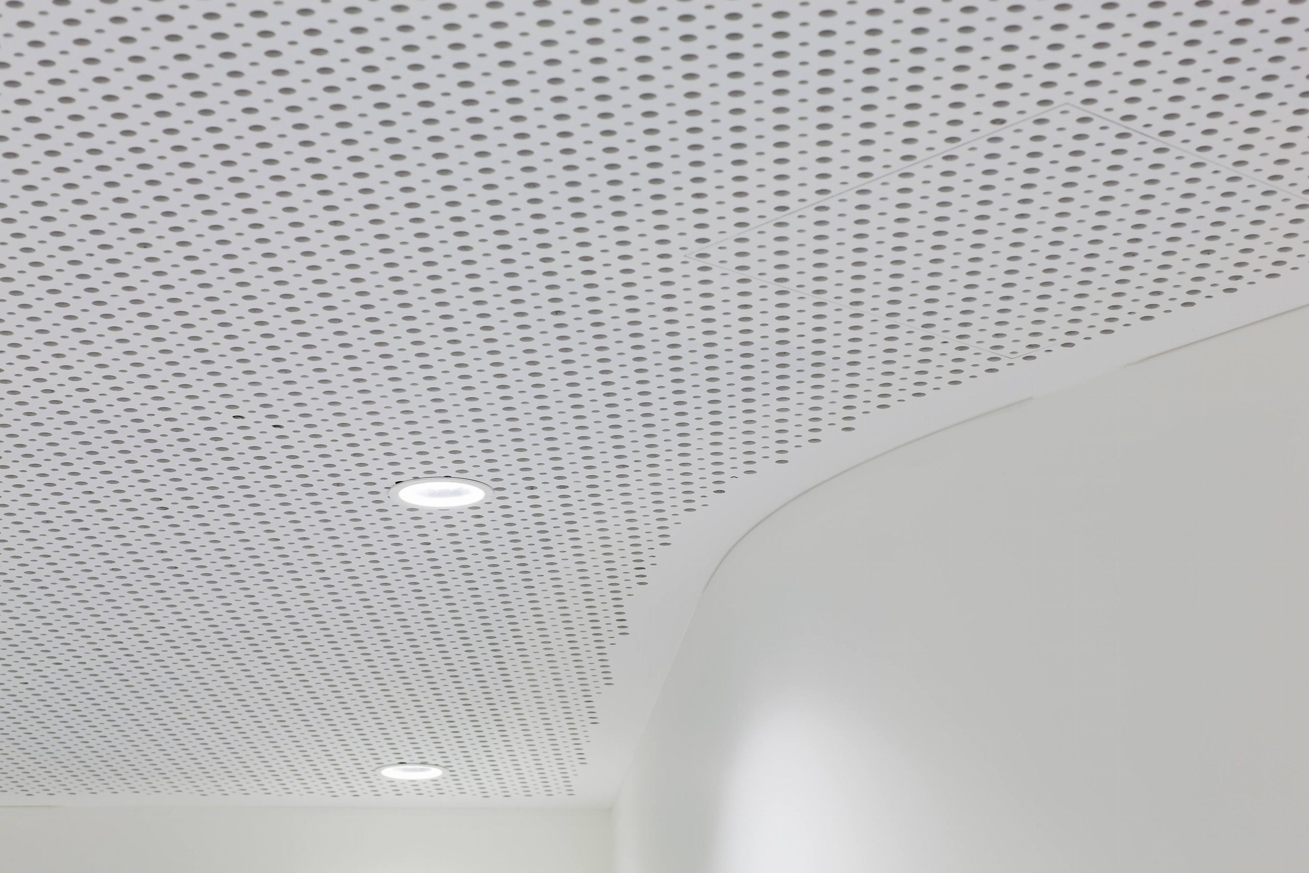 A white perforated ceiling with a curved edge and two round recessed lights. The perforations form a regular pattern across the ceiling, and part of a smooth white wall is visible below.