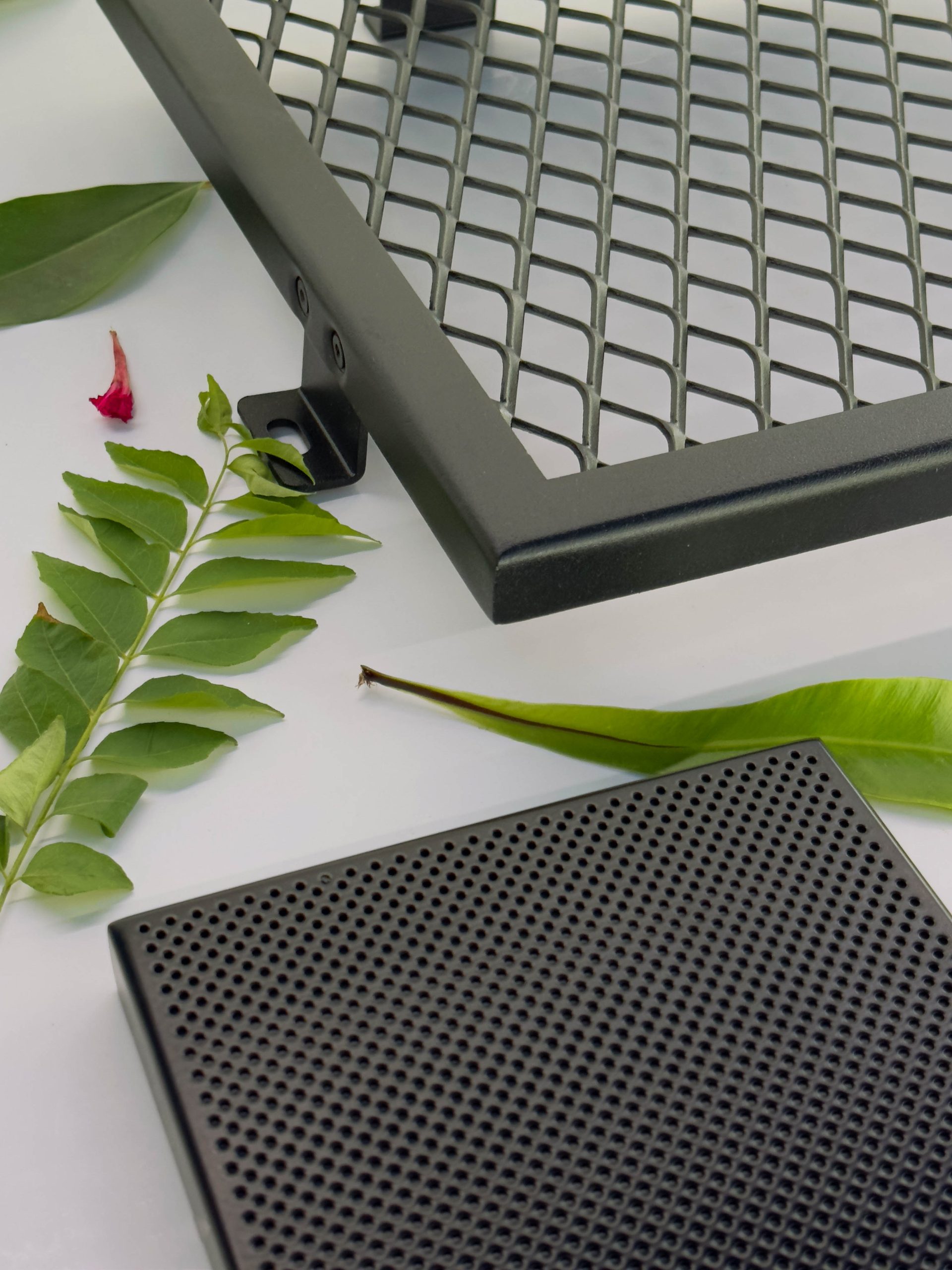 Several green leaves are arranged on a white surface next to a metal grid and a black perforated rectangular object, possibly electronic or automotive components.