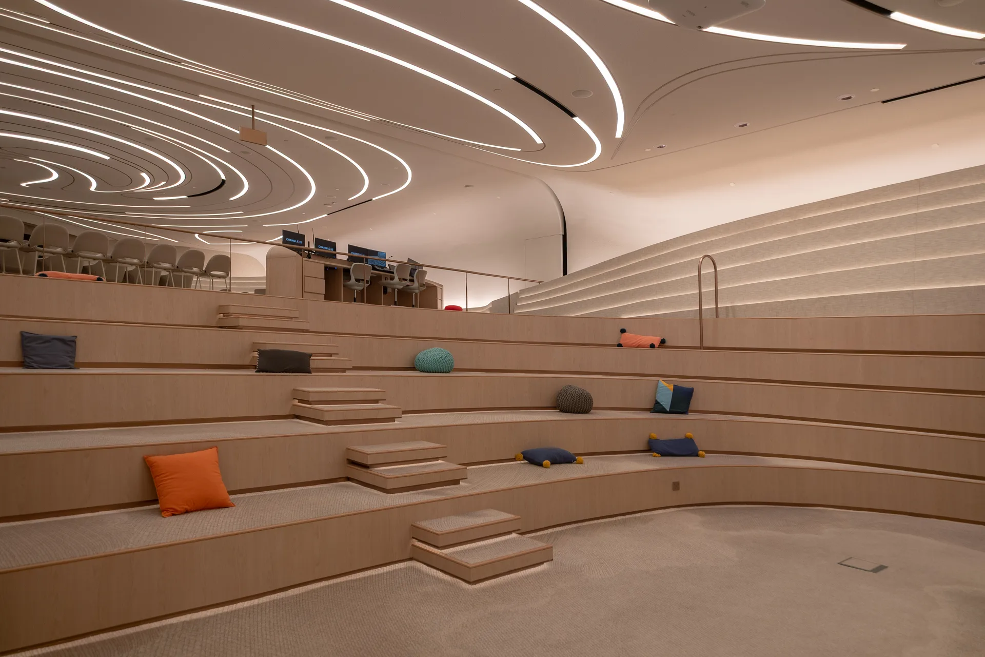 Modern, curved auditorium with wooden tiered seating, scattered colorful pillows, steps leading up, and a wavy, illuminated ceiling design. A control desk with monitors sits at the back near empty chairs.