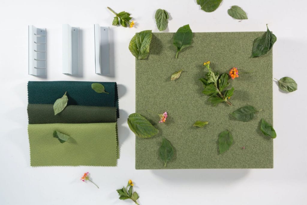 A green fabric square, scattered with leaves and small flowers, sits beside three rectangular gray samples and two fabric swatches—one dark green, one light green—on a white background.
