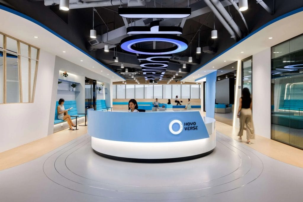 A modern, open office lobby with a curved blue and white reception desk, overhead circular lights, and people working or walking in a bright, spacious environment. The Hovo Verse logo is displayed on the desk.