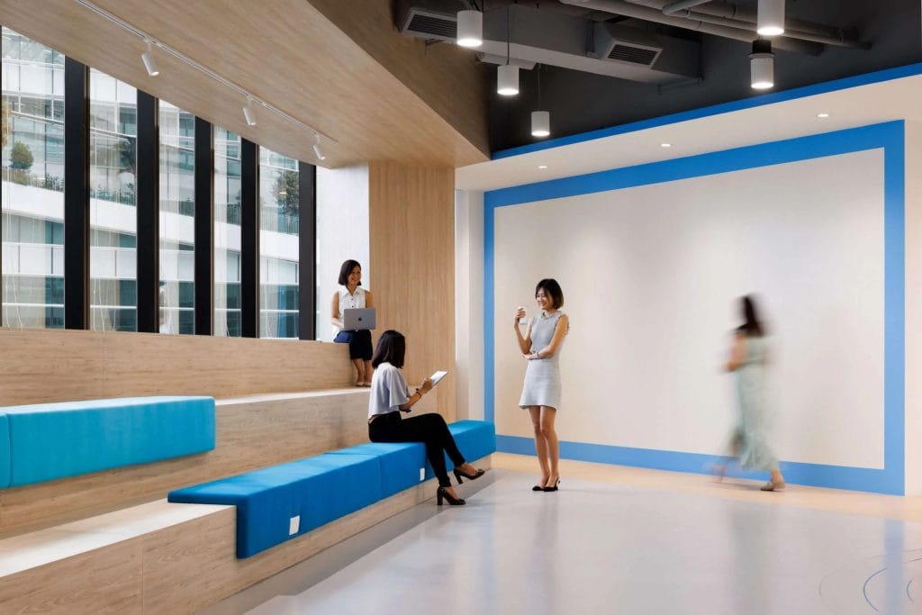 Modern office lounge with tiered wooden seating and blue cushions; three women are seated and talking, while another walks by, with large windows and bright, minimalist decor.