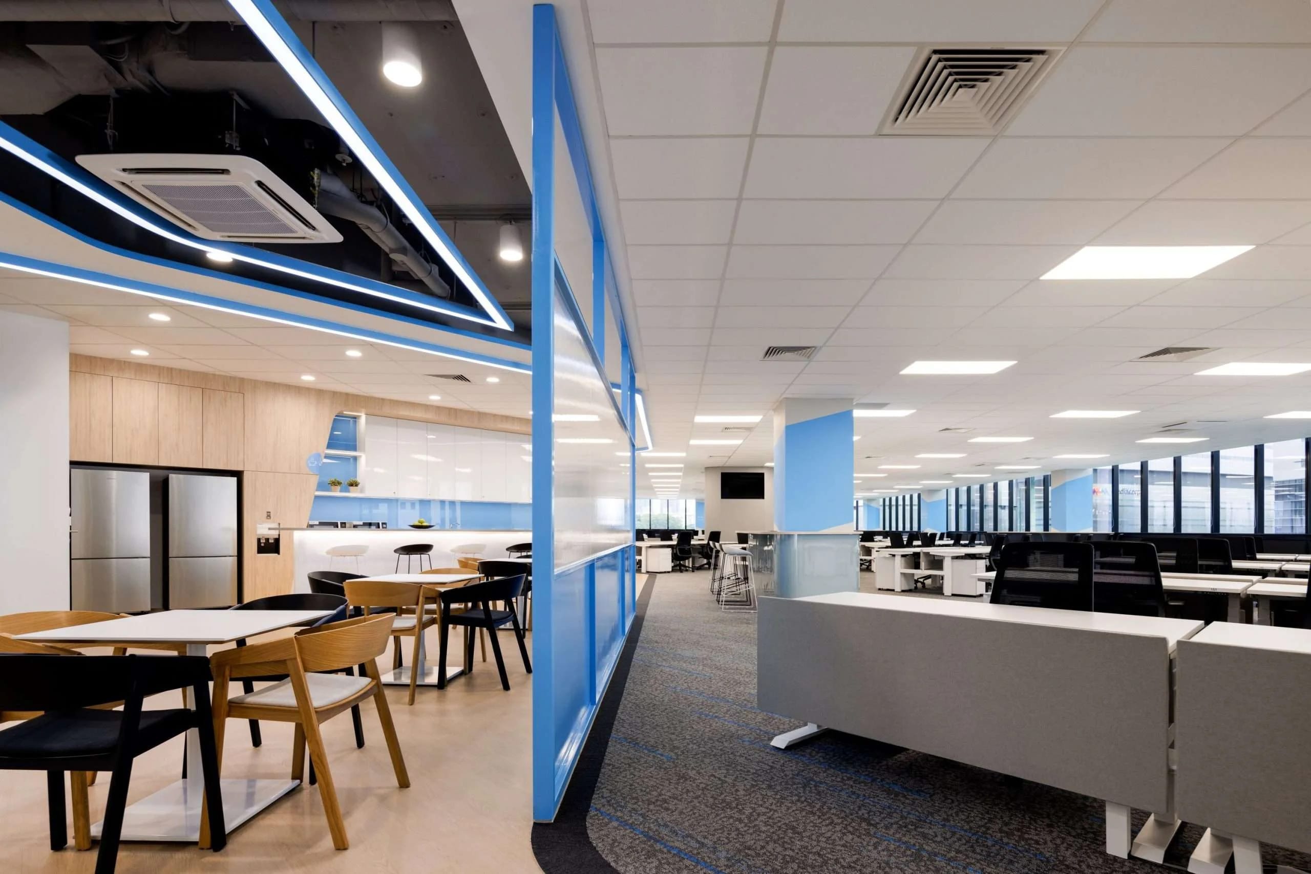 Modern open-plan office with rows of desks and computers, a glass partition, and a break area featuring wooden tables and chairs. The space is bright with ceiling lights and large windows along one wall.