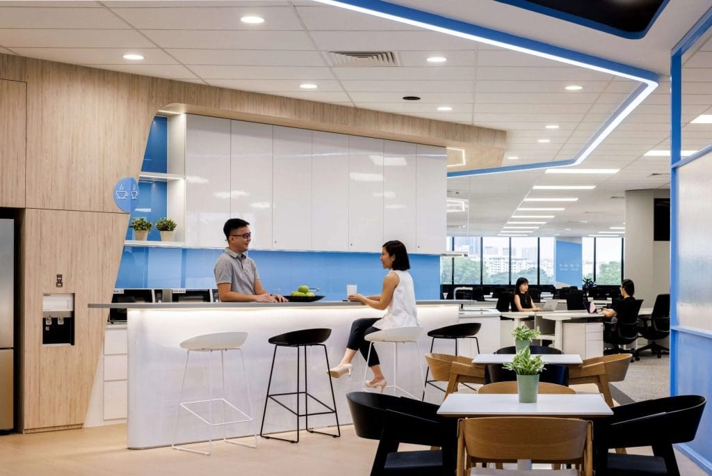 Modern office kitchen and lounge area with two people talking at a counter, surrounded by white cabinets, blue accent walls, and seating. Other employees are working in the background at desks.