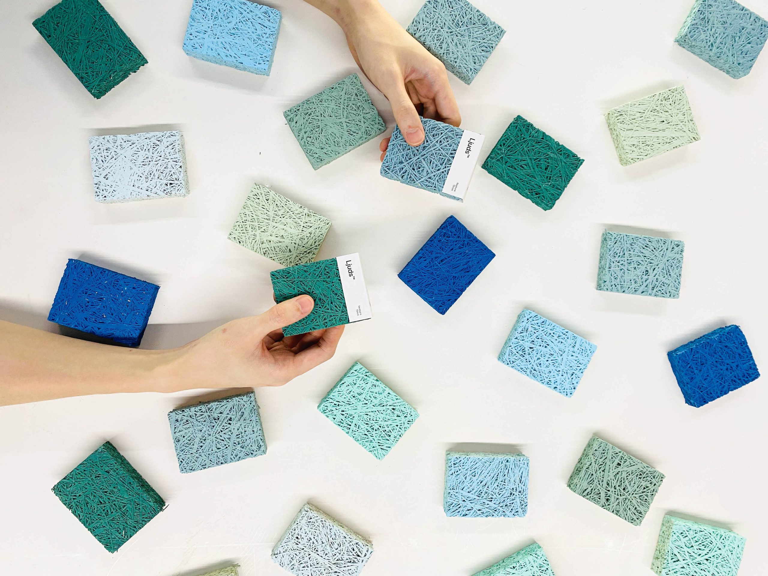 Two hands hold textured, rectangular color samples above a surface scattered with similar swatches in shades of blue, teal, and light green. The samples have a fibrous, woven appearance.