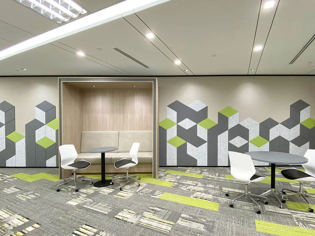 Modern office space with geometric wall designs, a built-in seating nook, round tables, and white chairs on wheels. The decor features gray and green tones with patterned carpet and bright lighting.