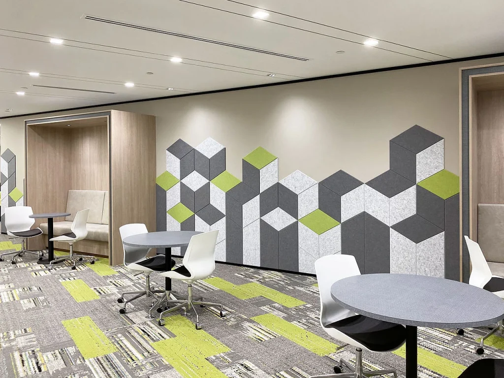 A modern office lounge with round tables, white chairs on wheels, and built-in seating alcoves. The wall features geometric gray, white, and green panels. The carpet has a matching green and gray pattern.
