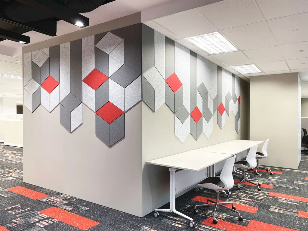Modern office space with white desks and chairs along a wall decorated with geometric, three-dimensional gray and red panels. The floor has a matching pattern with red and gray accents.