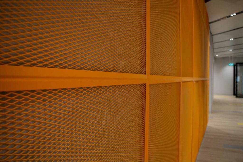 A close-up view of an orange metal mesh wall with a geometric pattern in a modern interior space, with a wooden floor and ceiling lights visible in the background.