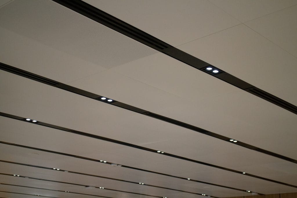 A modern ceiling with rows of recessed strip lighting and spotlights, creating a sleek, linear pattern. The panels are smooth and white, giving a clean, contemporary look.