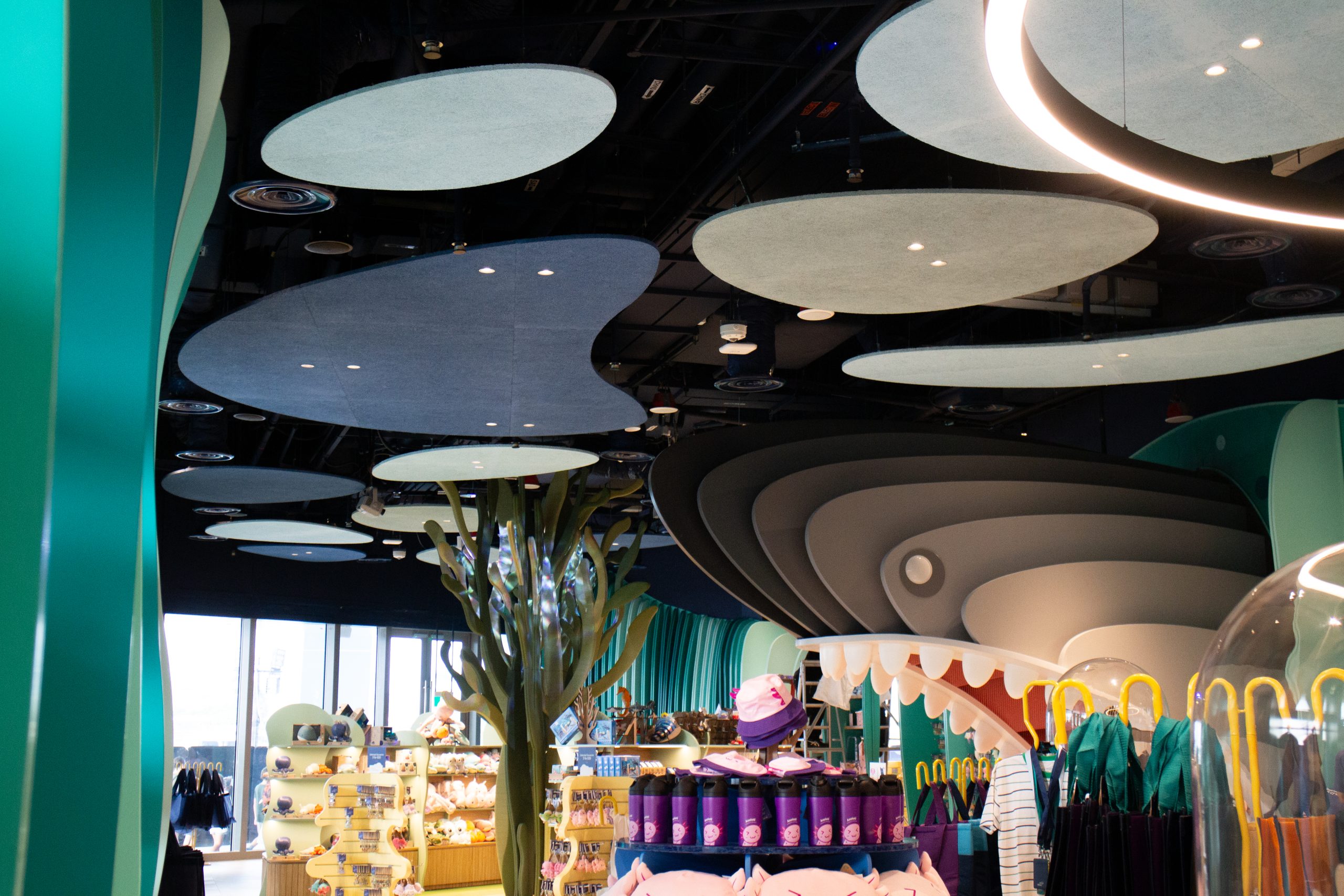 A whimsical retail store interior with wavy ceiling panels, teal columns, merchandise displays with colorful plush toys, hats, and clothing, and bright natural light from large windows.