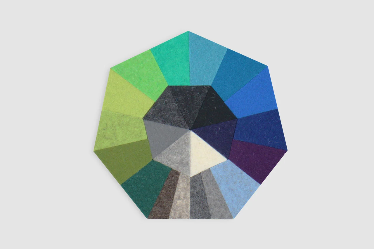 A circular arrangement of colored felt segments, each in a different shade, forming a color wheel against a light gray background. The colors range from greens and blues to grays and purples.