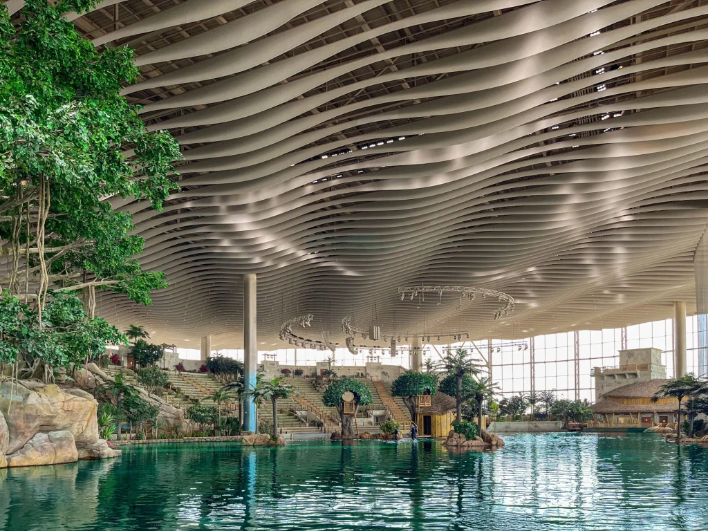Indoor tropical pool with lush greenery, artificial rocks, and small huts, surrounded by clear water. The wavy, modern ceiling and large windows let in natural light, creating a spacious and relaxing atmosphere.