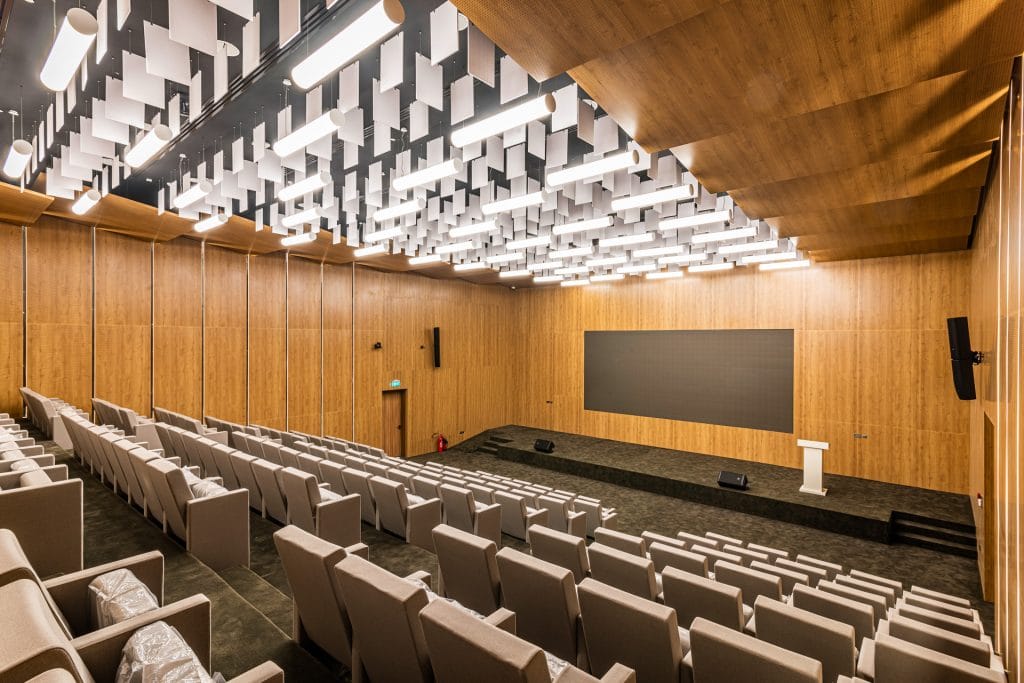 A modern lecture hall with tiered gray seating, wood-paneled walls, a large screen at the front, a white podium, and a ceiling featuring white acoustic panels and bright lighting.