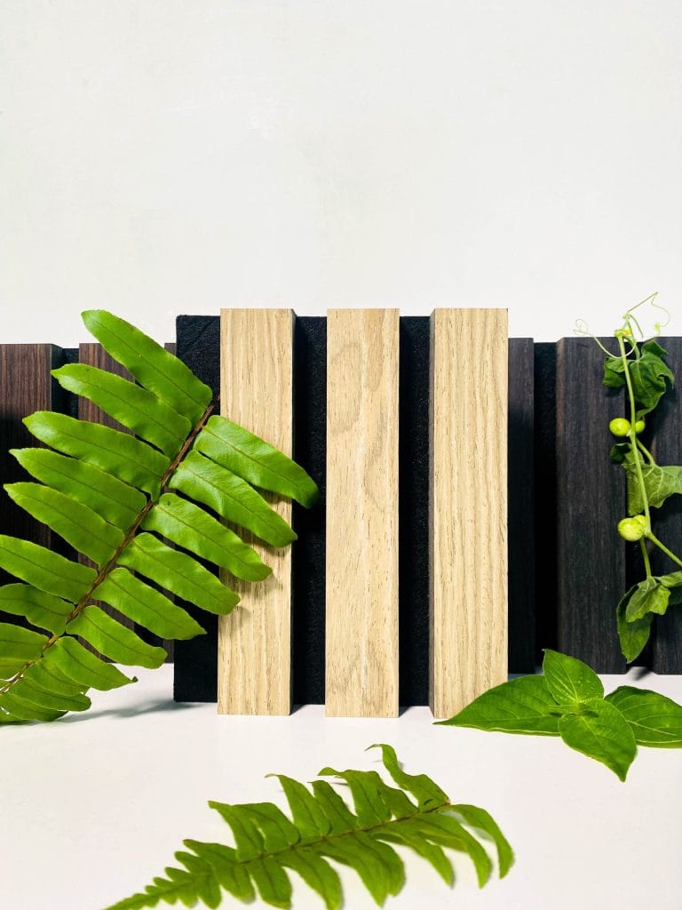 Three light wooden blocks stand vertically between dark wooden planks, with green fern leaves and small vines artfully arranged in the foreground and background against a white backdrop.