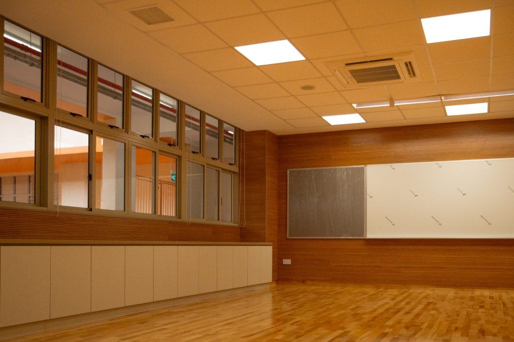 A well-lit, empty classroom with wooden floors, large windows, a whiteboard, and wooden panel walls. The ceiling has recessed lighting and an air conditioning unit.