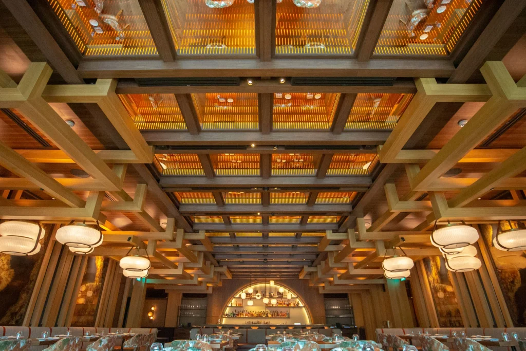 Elegant restaurant interior with a high wooden lattice ceiling, large round hanging lights, and a well-lit bar at the back. Warm lighting and carefully set tables create a sophisticated, welcoming atmosphere.