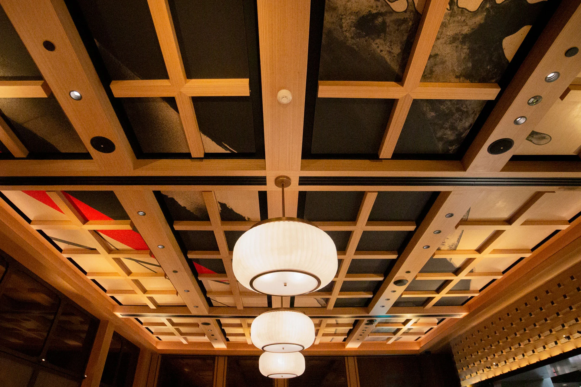 A wooden coffered ceiling with three large, round, white pendant lights hanging in a row. The ceiling panels display abstract art, and spotlights are embedded between the grids.