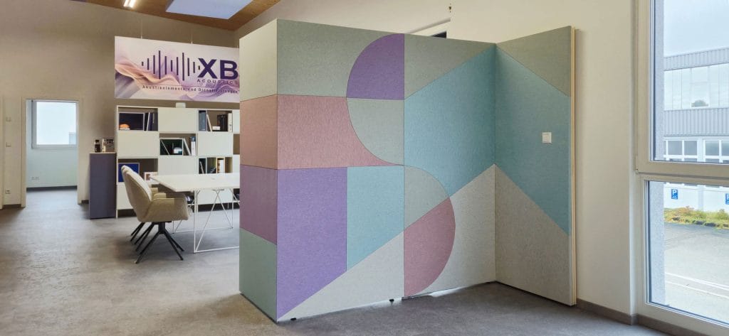 A modern office with a pastel geometric-patterned privacy screen, white shelving with books, beige chairs around a table, and large windows letting in natural light.