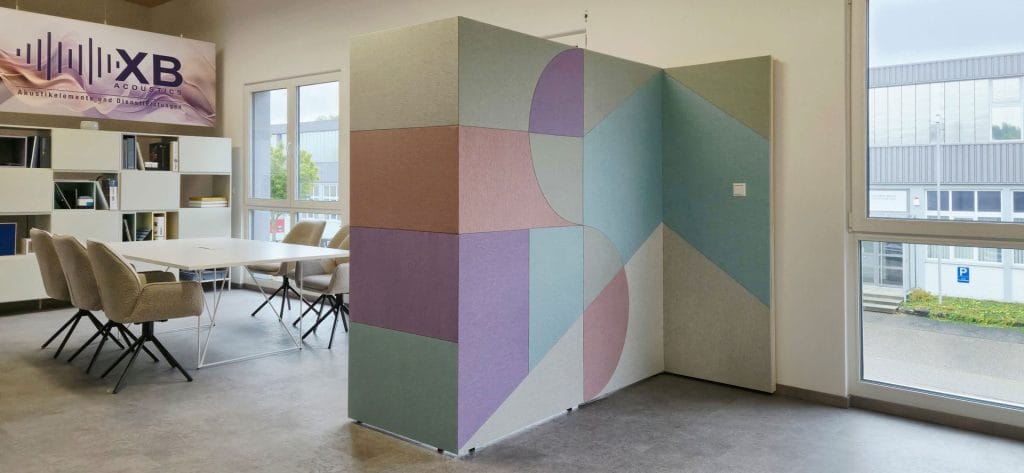 A modern office space with a geometric-patterned, pastel-colored room divider near a table with beige chairs, bookshelves, and large windows letting in natural light.