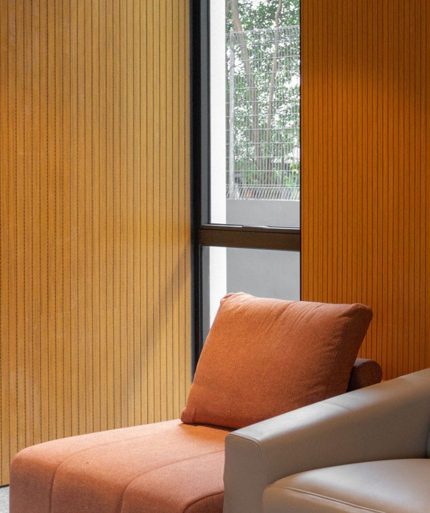 A cozy corner with an orange cushioned chair and part of a light gray sofa, set against vertical wood paneling and a window showing greenery outside.