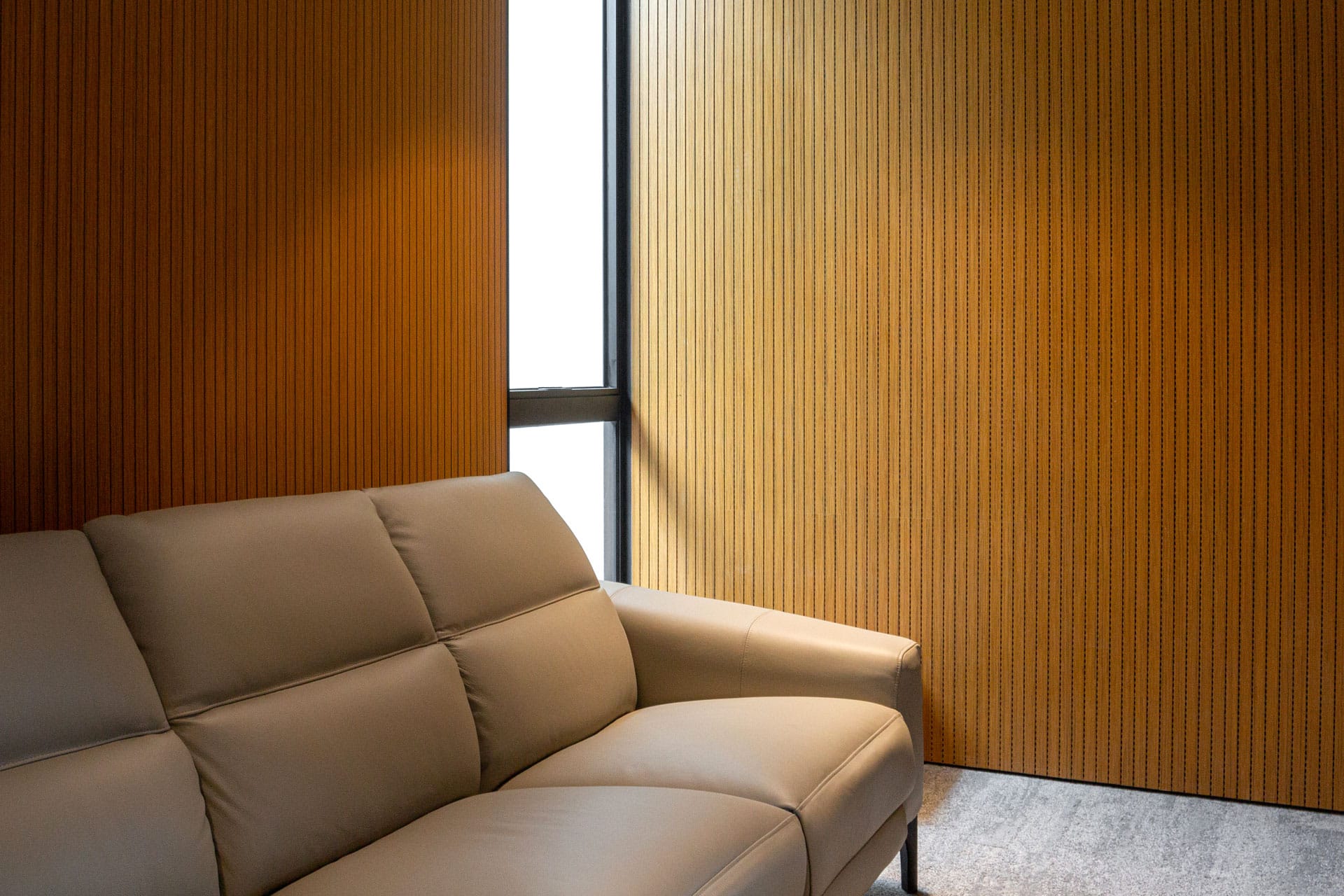 A beige leather sofa sits in front of wood-paneled walls with vertical grooves and a narrow window letting in natural light. The setting appears modern and minimalistic.