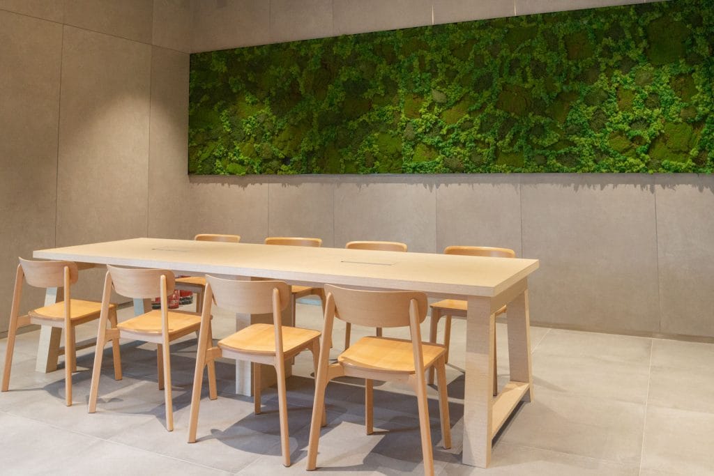 A modern meeting room with a long light wooden table, eight matching wooden chairs, beige tiled walls and floor, and a large green moss wall art piece as decoration.