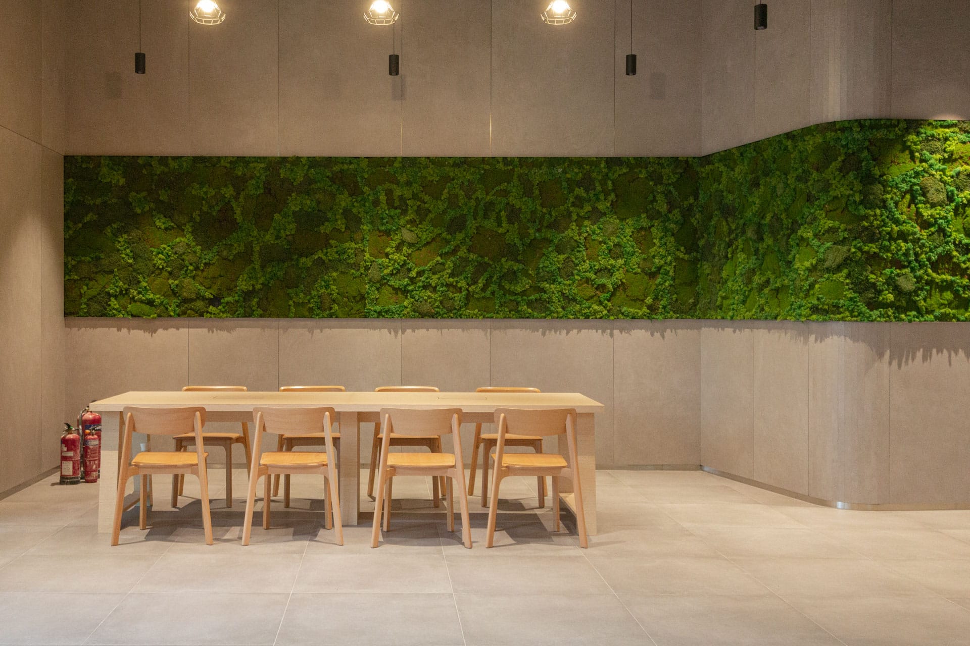 A modern dining area with a light wooden table and six matching chairs, set against a gray wall featuring a large panel of lush green moss and three hanging pendant lights above.