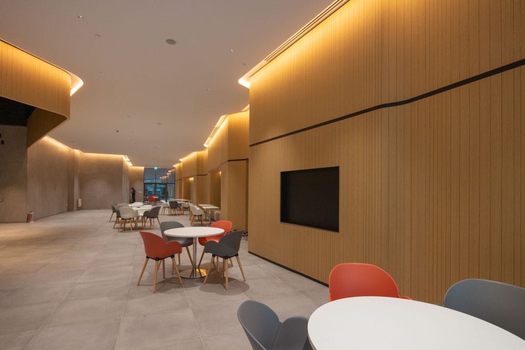 A modern lounge area with round tables and colorful chairs, light wood-paneled walls, soft ceiling lighting, and large windows in the background. The space appears empty and spacious.