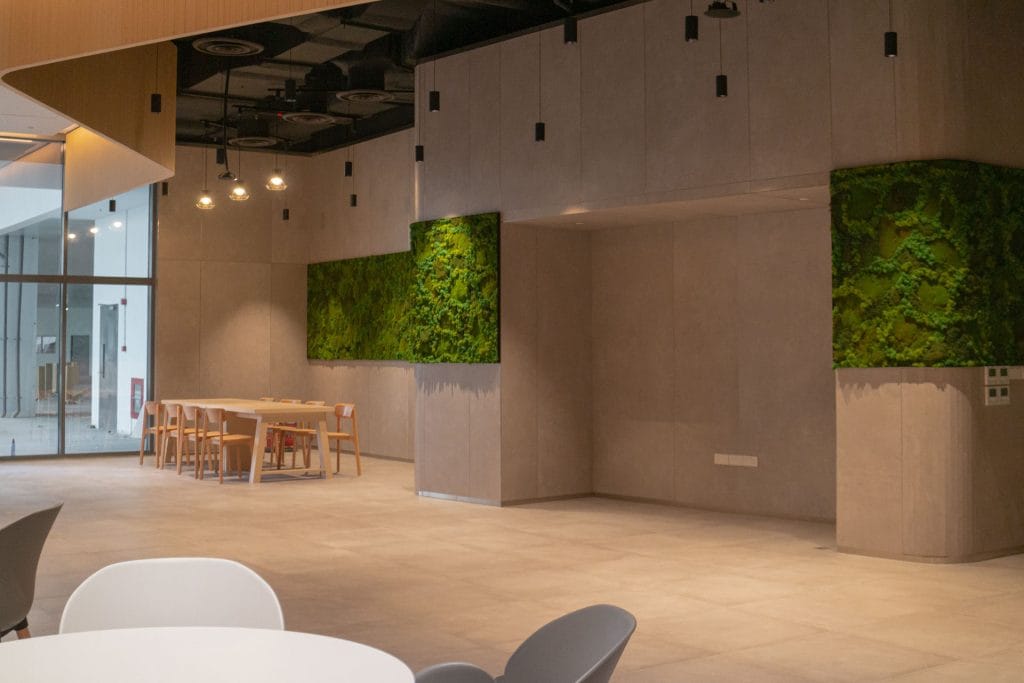 A modern, minimalistic office space with light wood furniture, beige tiled floors, green vertical plant panels on the walls, and large windows letting in natural light.