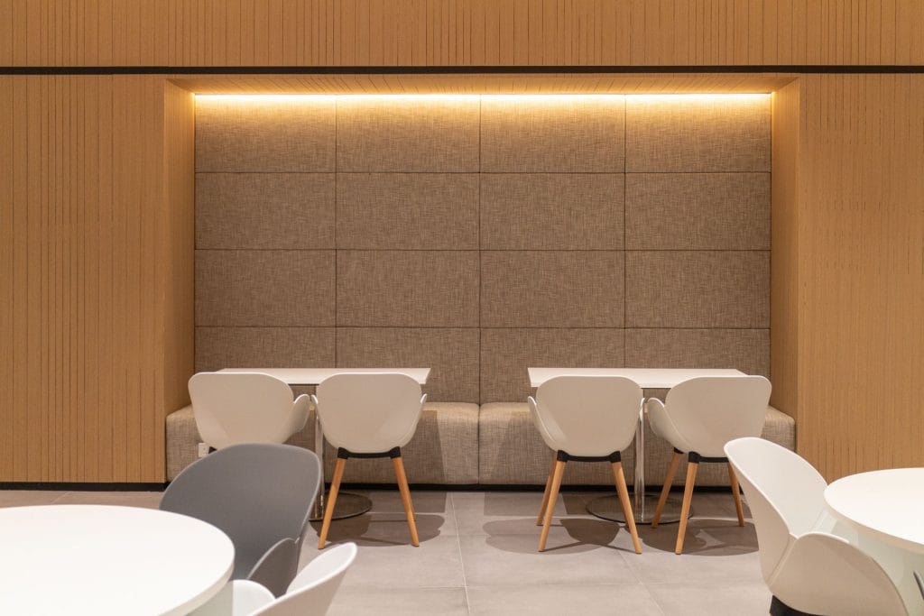 A modern, minimalistic dining area with white chairs and round tables. The background features a padded wall with beige upholstery, wood paneling, and soft, warm overhead lighting. The space appears clean and well-organized.