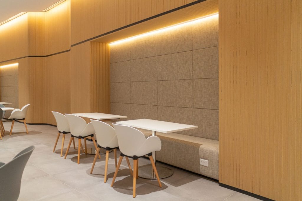 A modern, minimalistic dining area with light wood walls, white tables, and white chairs with wooden legs. A built-in cushioned bench runs along the wall under warm recessed lighting.