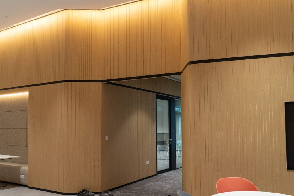 A modern office interior with wooden panel walls, recessed ceiling lighting, a glass door leading to another room, and a corner with round tables and colorful chairs in the foreground.