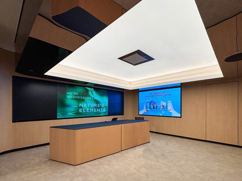 A modern conference room with a wooden podium, large screens displaying presentations, and sleek lighting. One screen reads, “We’re harnessing nature’s elements,” while the other shows lab equipment.