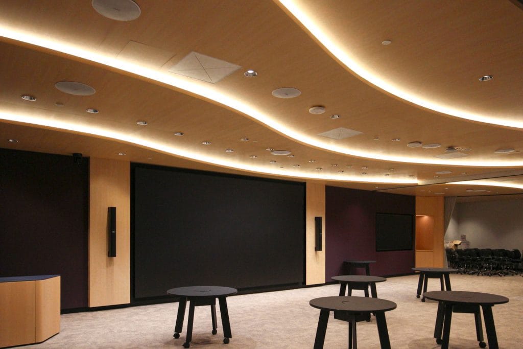 A modern conference room with curved, illuminated ceiling panels, a large blank screen on the wall, circular tables without chairs, and additional seating arranged in rows at the back.