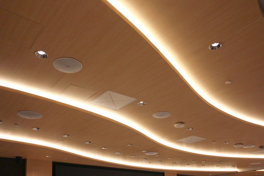 Curved wooden auditorium ceiling with built-in lights, speakers, and acoustic panels, illuminated by strips of recessed lighting.