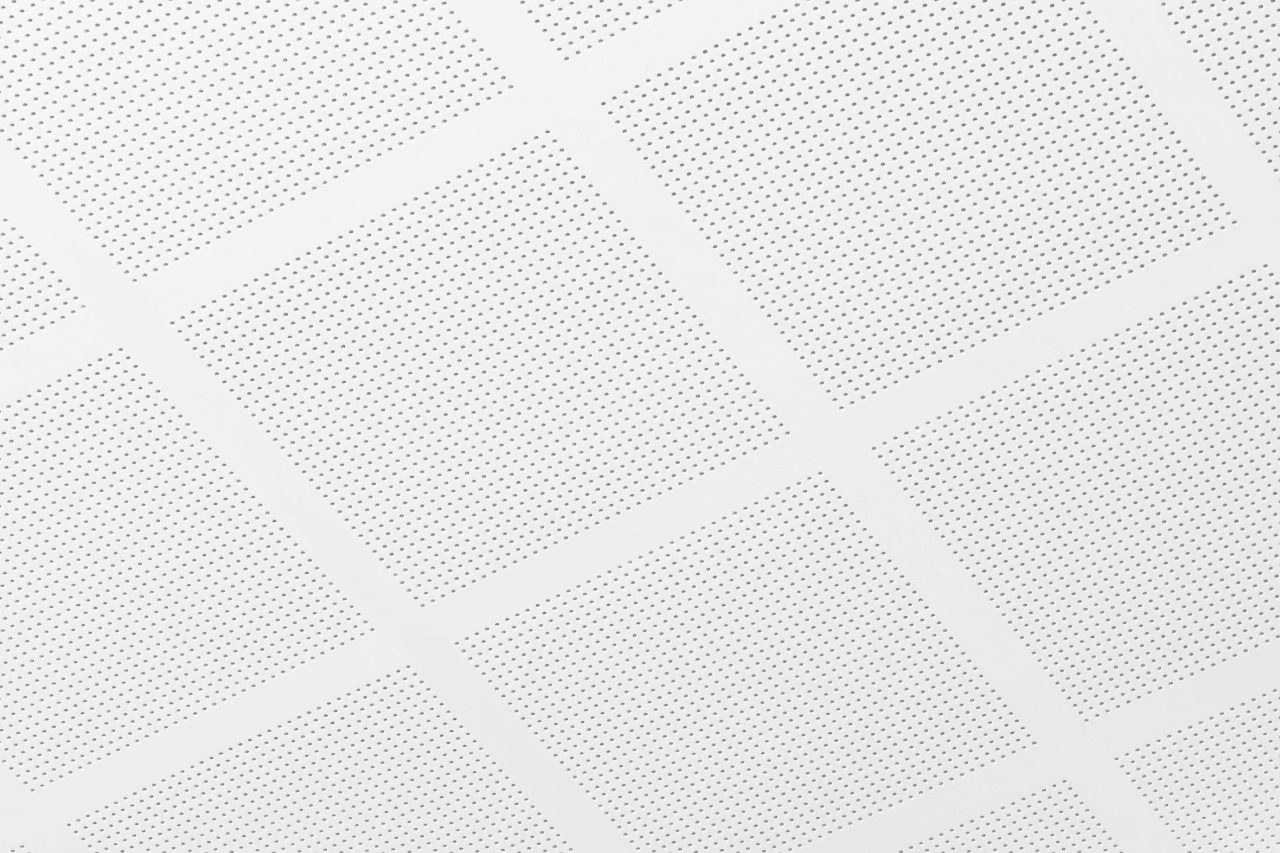 White ceiling panels with a grid pattern, each panel perforated with small holes, creating a uniform, geometric design. The image is well-lit and shows a close-up of the ceiling tiles.