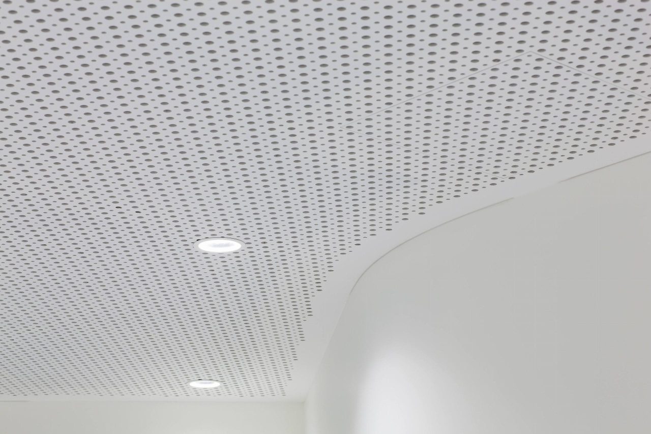 A white perforated ceiling with a curved edge and two round recessed lights. The perforations form a regular pattern across the ceiling, and part of a smooth white wall is visible below.