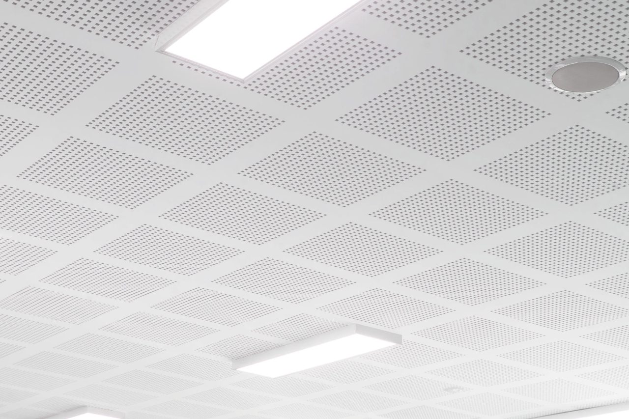 A white ceiling with a grid of perforated square panels, circular speaker, and rectangular fluorescent lights embedded in the surface. The overall appearance is clean and modern.