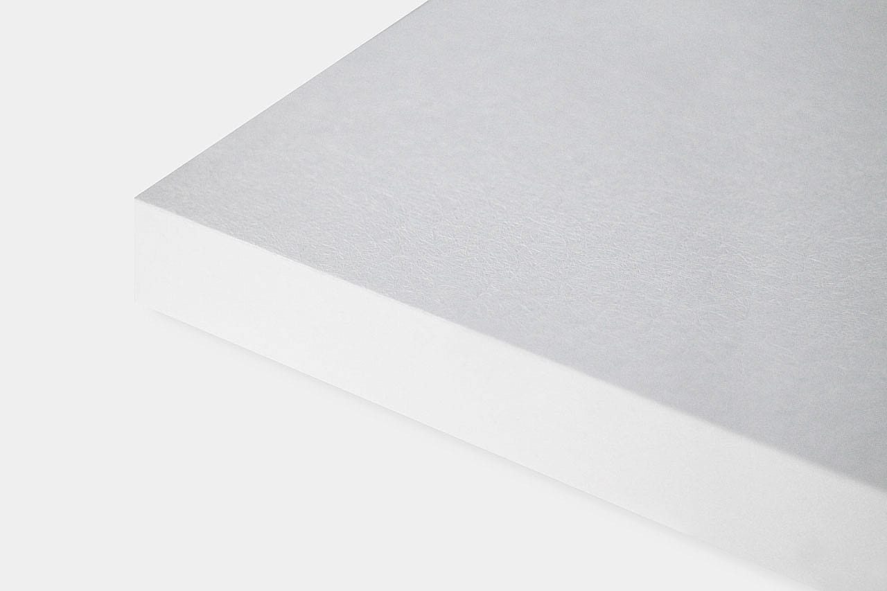 A close-up of the corner of a white, rectangular sheet of paper or cardstock on a plain light background. The surface shows a subtle, textured finish.